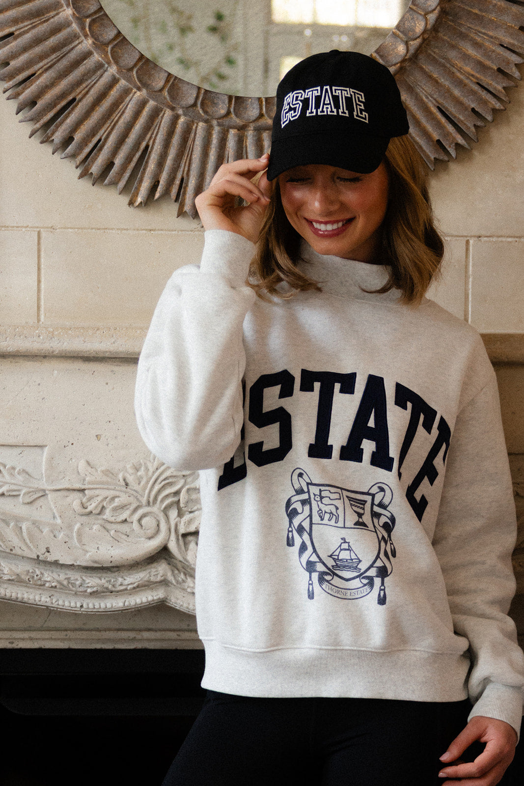 Person wearing a 'ESTATE' sweatshirt and Estate cap in front of a decorative mirror.