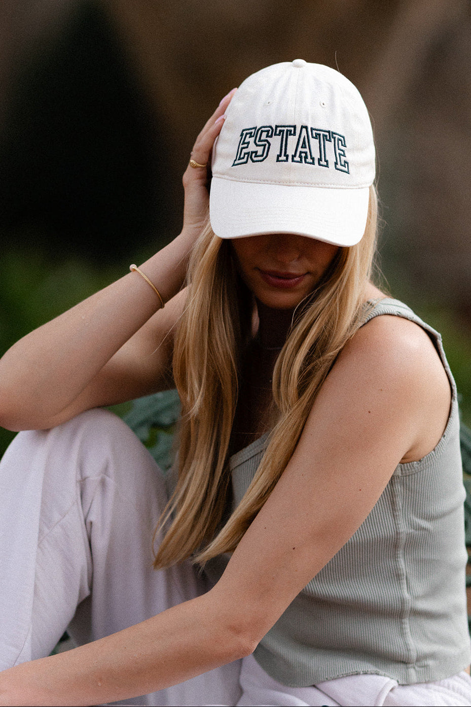 Woman wearing a white cap with 'ESTATE' embroidery sitting outdoors.